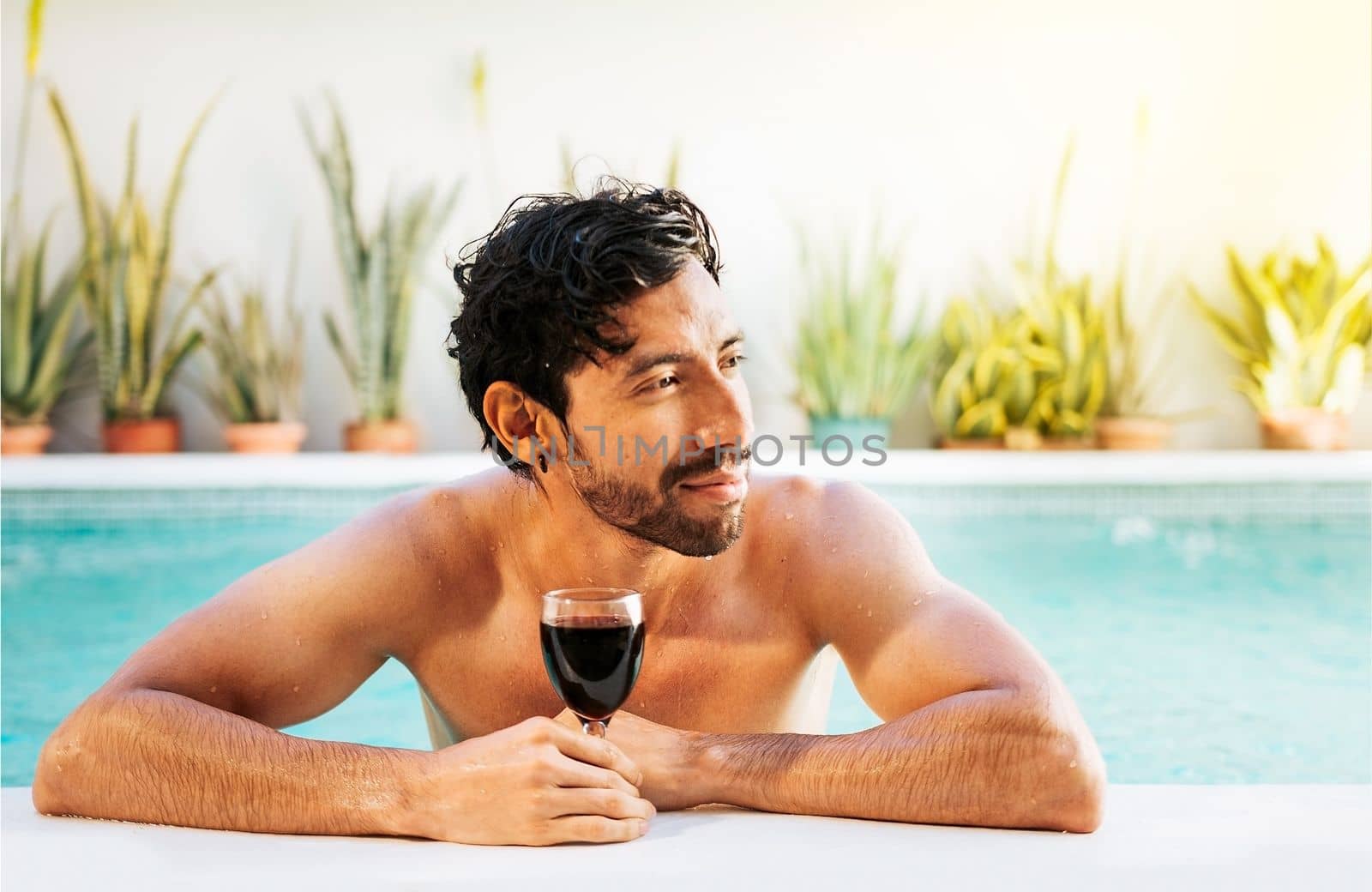 Portrait of man at the edge of swimming pool with a glass of wine. Handsome man in swimming pool holding glass of wine. Handsome guy on the edge of the pool holding a glass of wine