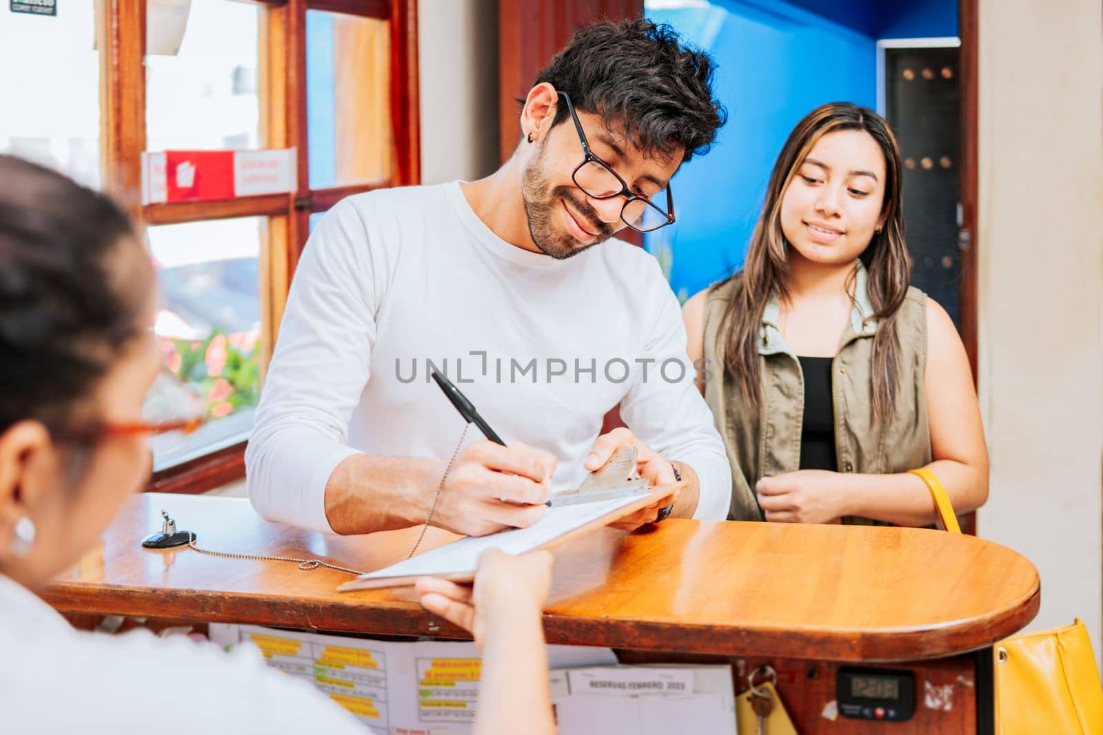 Couple signing in at a hotel reception. Young couple signing documents in a hotel reception. Latin couple signing reservation at hotel reception. Hotel reservation concept by isaiphoto