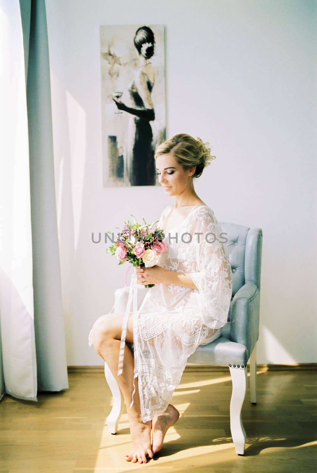 Bride in a lace dressing gown with a bouquet sits in a chair. High quality photo