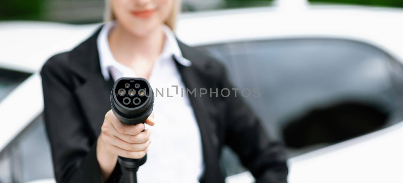 Blur closeup businesswoman hand holding and pointing an EV plug at camera for electric vehicle as progressive idea of alternative sustainable clean and green energy for environmental concern.