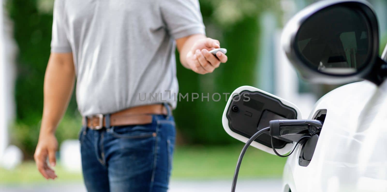 Progressive asian man install cable plug to his electric car with home charging station in the backyard. Concept use of electric vehicles in a progressive lifestyle contributes to clean environment.