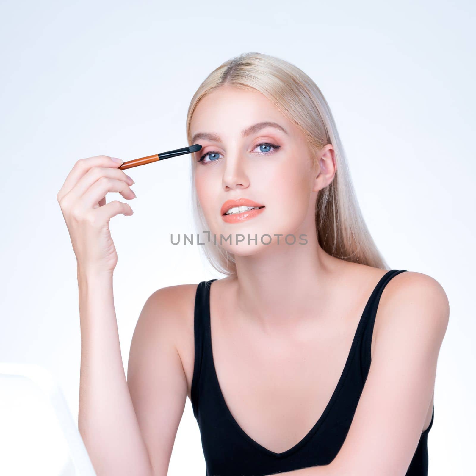 Beautiful personable girl with flawless applying eye shadow makeup with eyeliner brush. Cosmetic facial painting process on lovely young woman with perfect clean skin in isolated background.