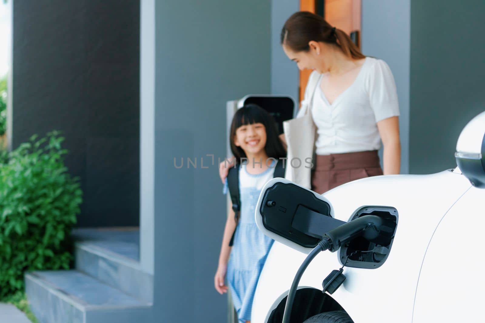 Focus electric vehicle recharging at home charging station plugged in with EV charger device with blurred background of progressive mother and daughter walking as concept for sustainability of energy.