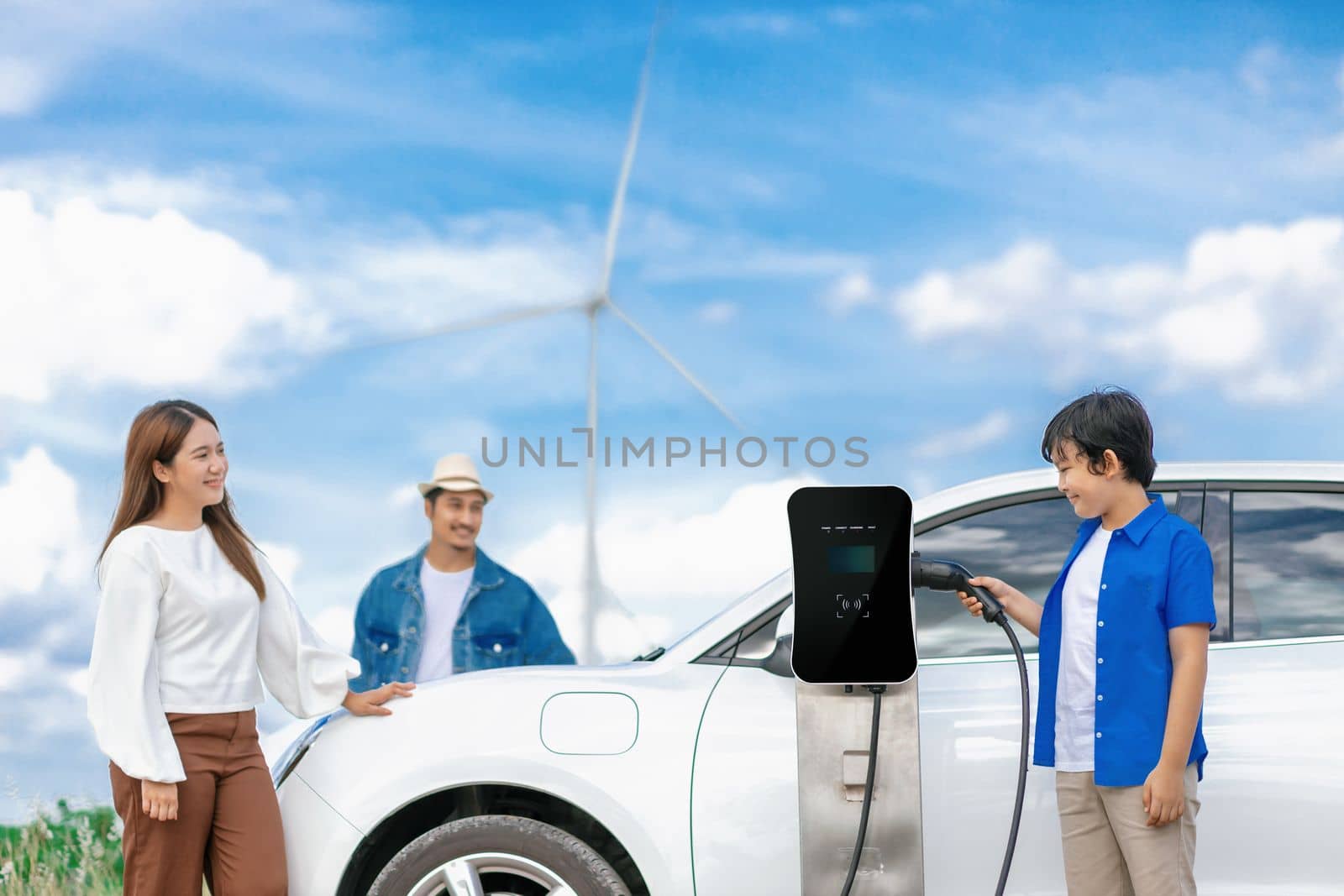 Concept of progressive happy family enjoying their time at wind farm with electric vehicle. Electric vehicle driven by clean renewable energy from wind turbine generator for charging station.