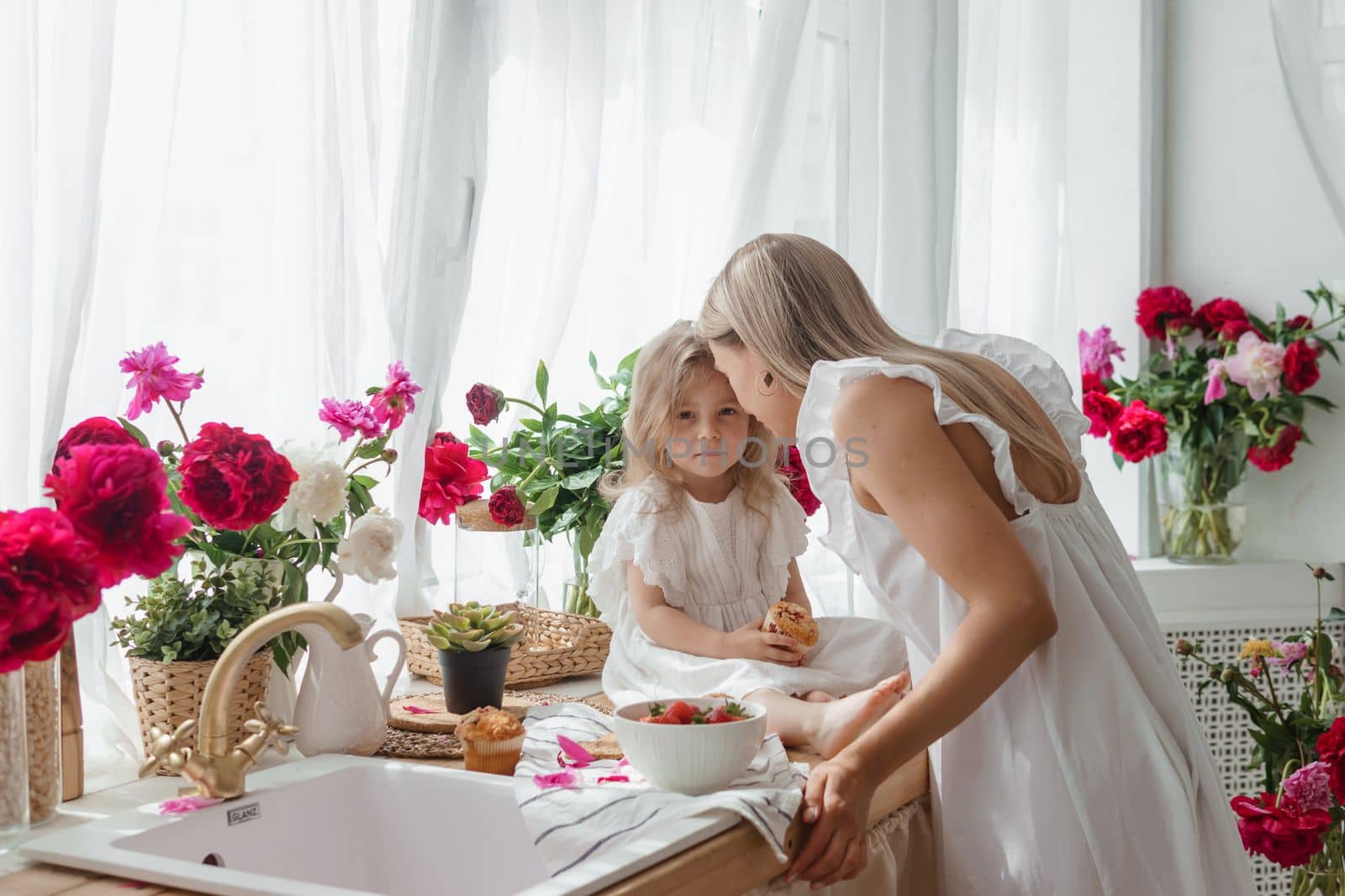 A little blonde girl with her mom on a kitchen countertop decorated with peonies. The concept of the relationship between mother and daughter. Spring atmosphere.