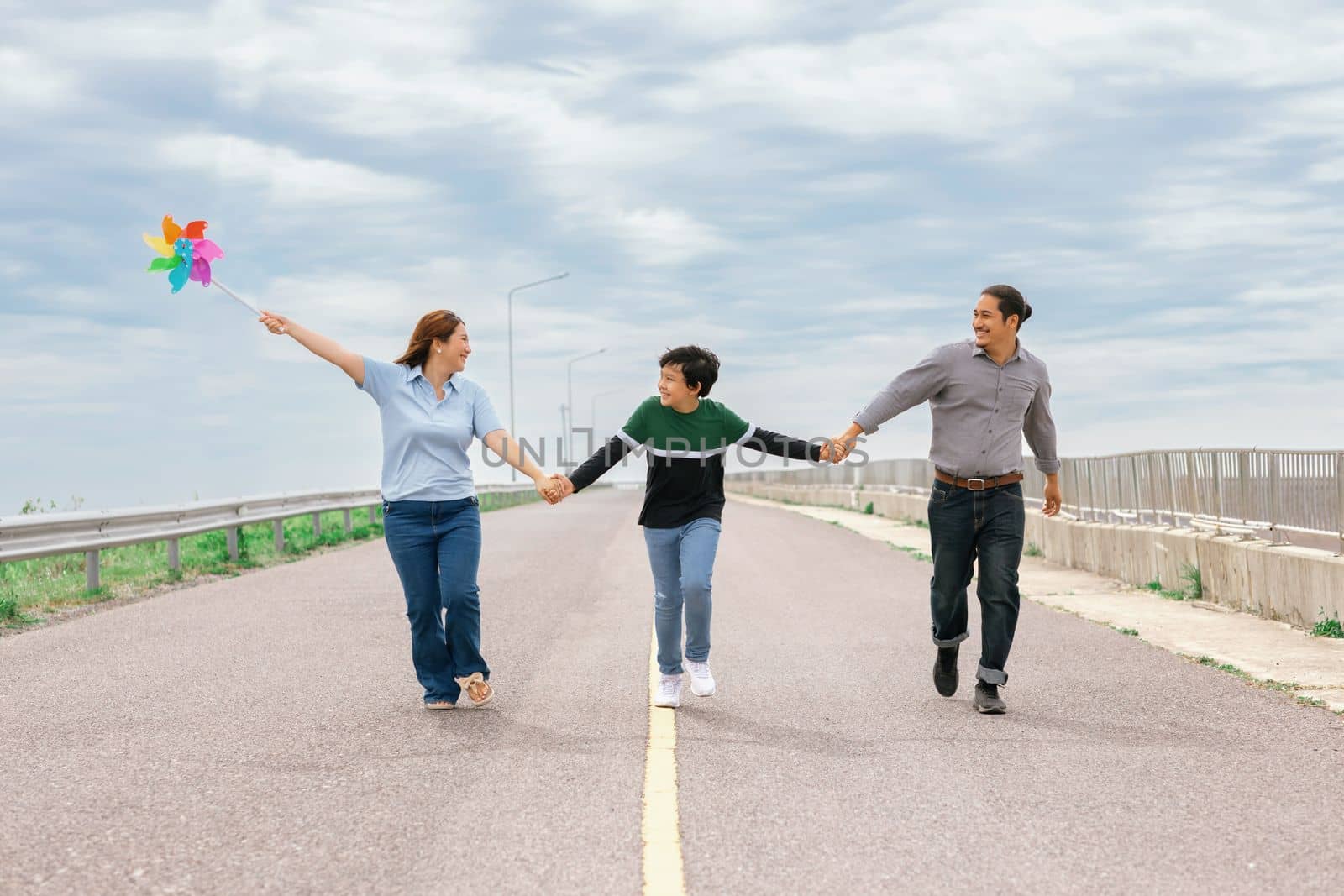 Progressive happy family vacation and carefree day concept. Young parents mother father and son run along and flying kite together road with enjoy natural scenic on scenery and clear sky background.