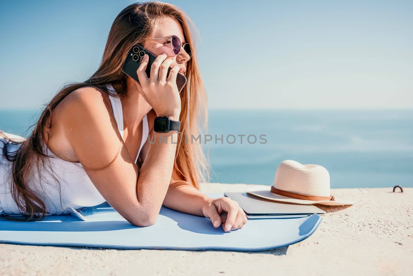 Digital nomad, woman in the hat, a business woman with a laptop sits on the rocks by the sea during sunset, makes a business transaction online from a distance. Freelance, remote work on vacation. by panophotograph