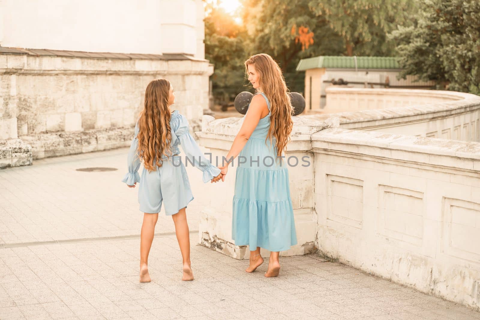 Daughter mother holding hands. In blue dresses with flowing long hair against the backdrop of a sunset and a white building