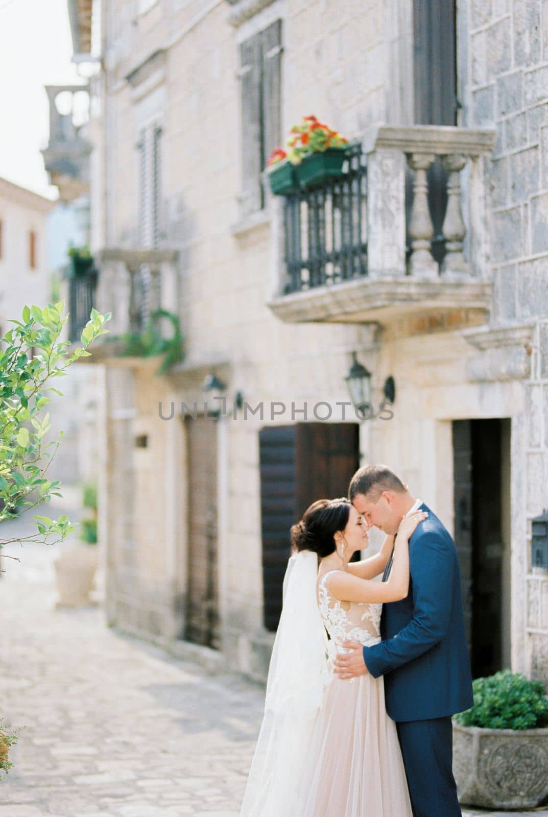 Groom hugs bride in a white dress near a stone building with balconies. High quality photo