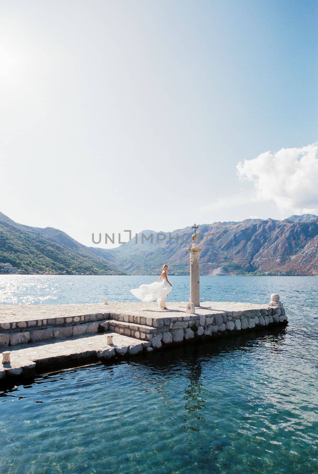 Bride in a flowing dress stands on a pier near a column with a cross. High quality photo