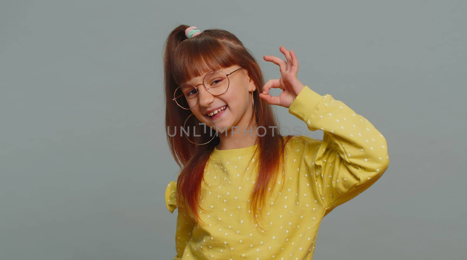 Ok. Young toddler school girl in glasses looking approvingly at camera showing okey gesture, like sign positive, approve something good. Teenager female child kid isolated on studio gray background
