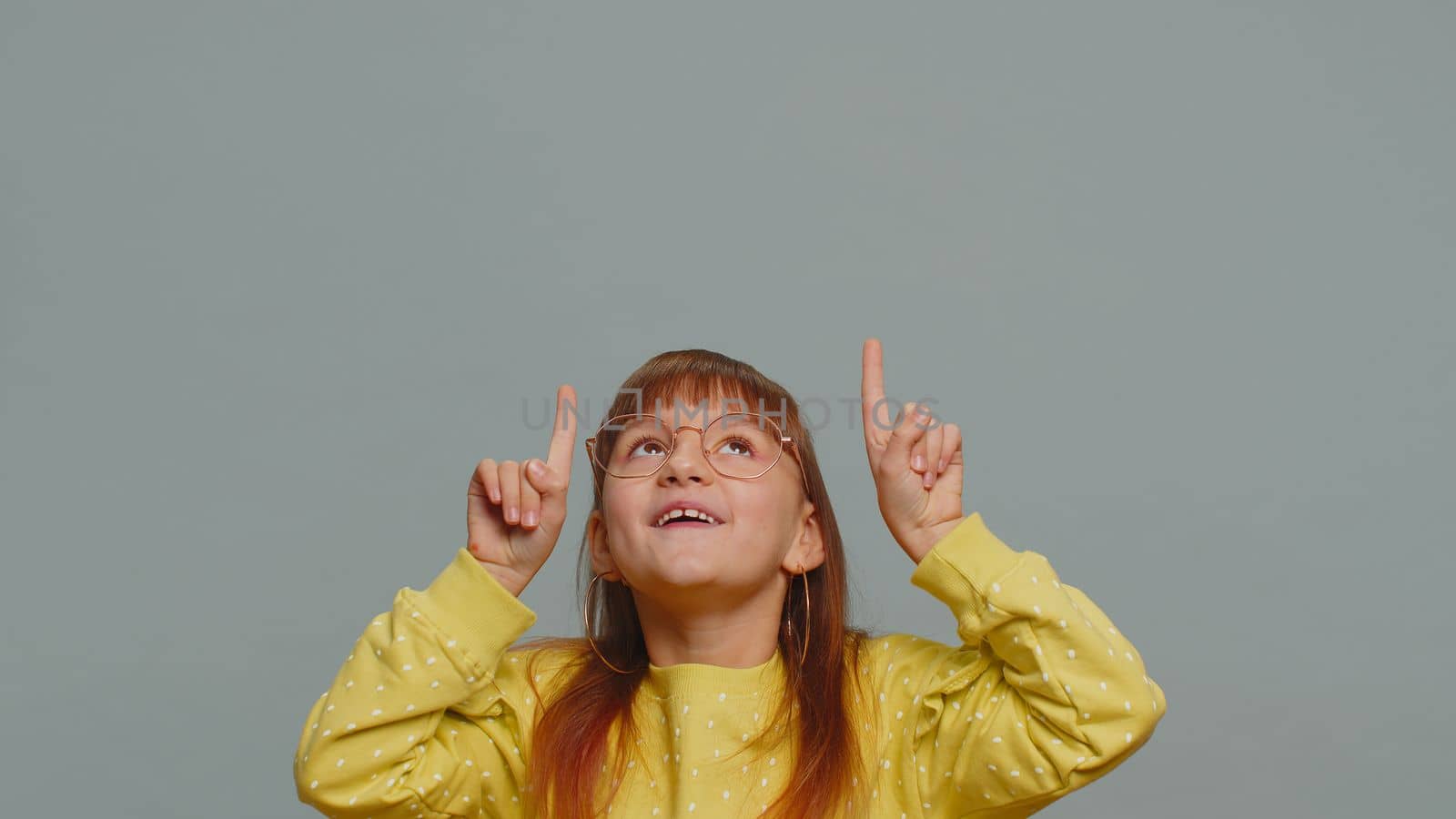 Happy toddler girl showing thumbs up pointing empty place up, advertising area for commercial text, copy space for goods promotion banner, copy-space. Young child kid. Studio shot on gray background