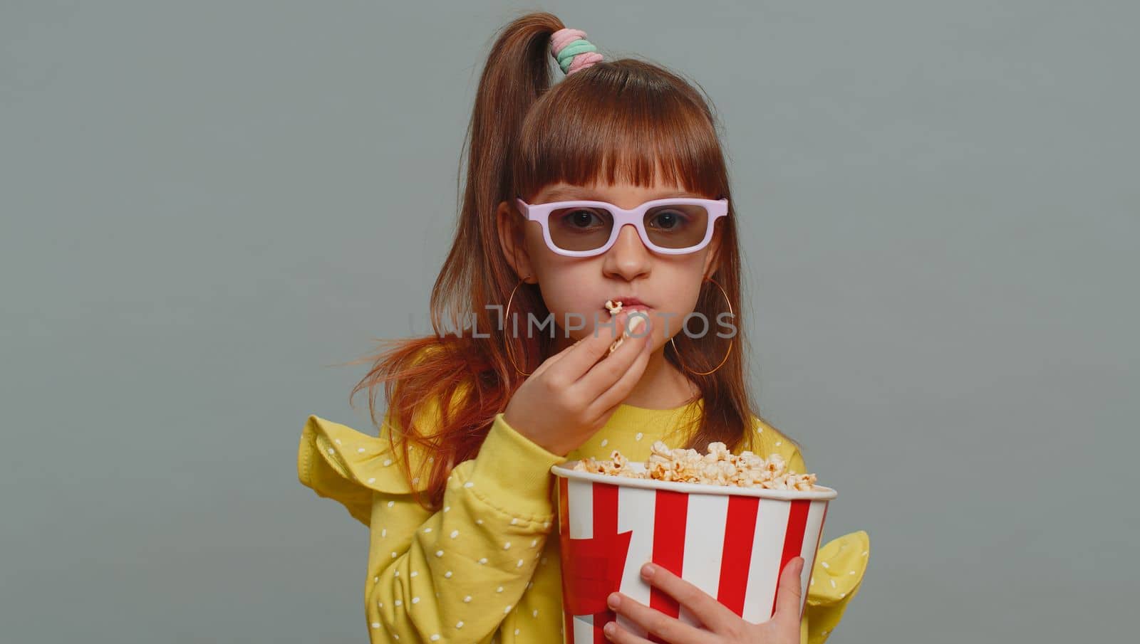 Excited young toddler school girl in 3D glasses eating popcorn, watching interesting tv serial, sport game, film, online social media movie content. Teen female child kid on studio gray background