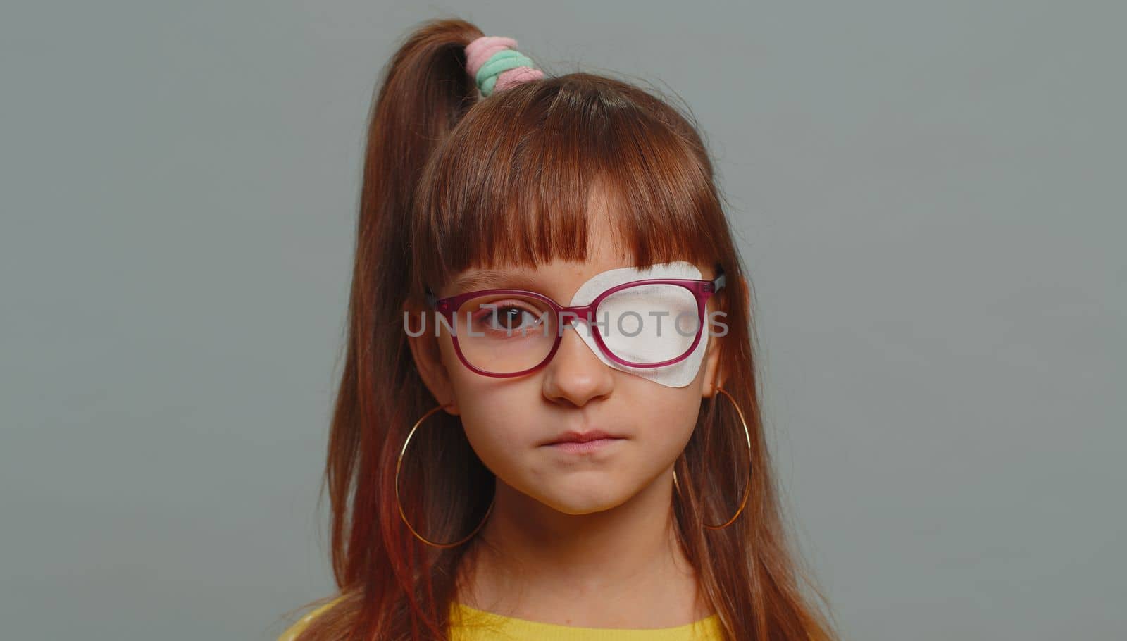 Sick ill preteen girl looking at camera having protective bandage plaster patch scratch on one eye. Injury, trauma, blind. Child kid wearing glasses at gray background. Medical eyesight vision therapy