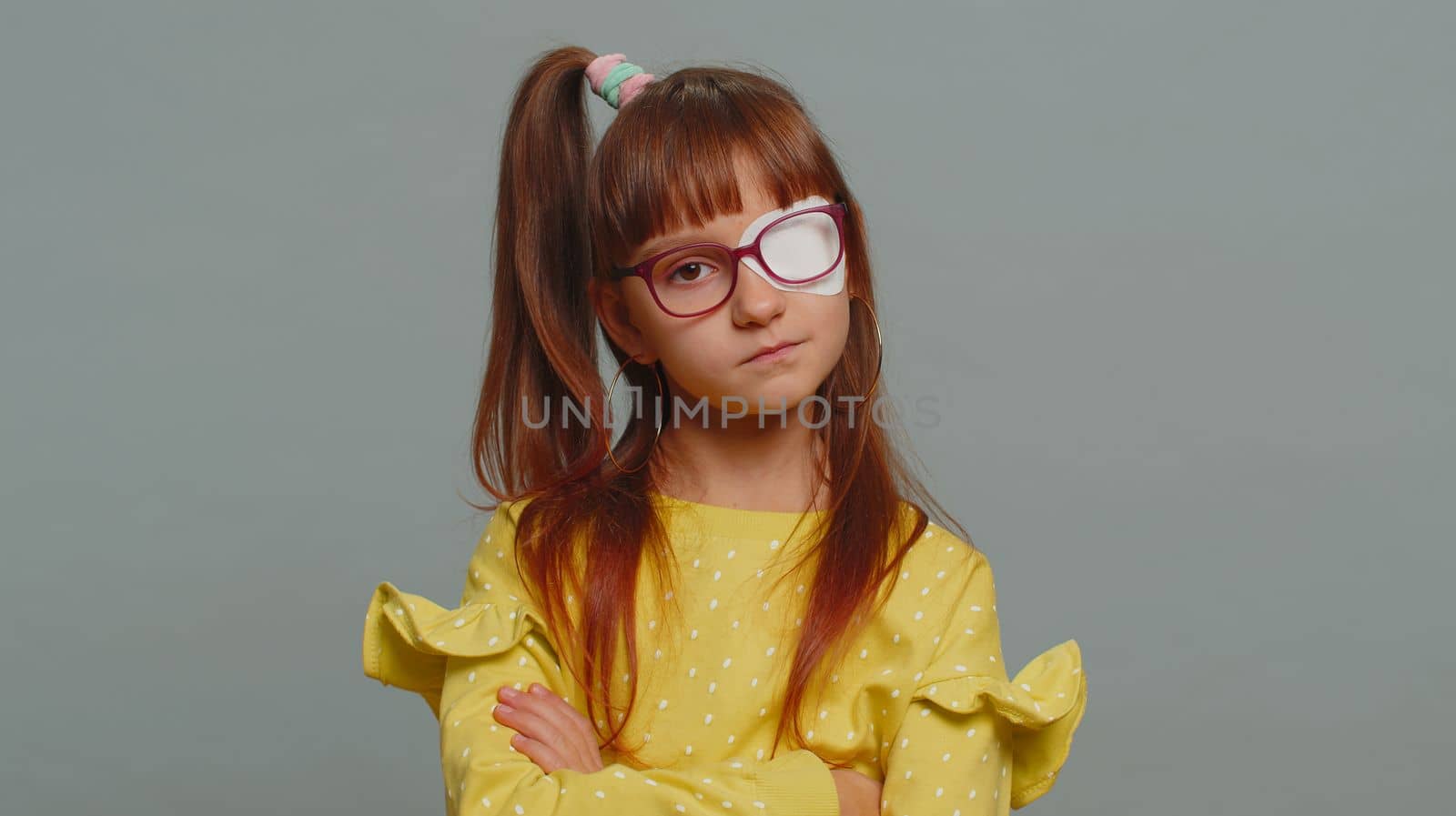 Sick ill preteen girl looking at camera having protective bandage plaster patch scratch on one eye. Injury, trauma, blind. Child kid wearing glasses at gray background. Medical eyesight vision therapy