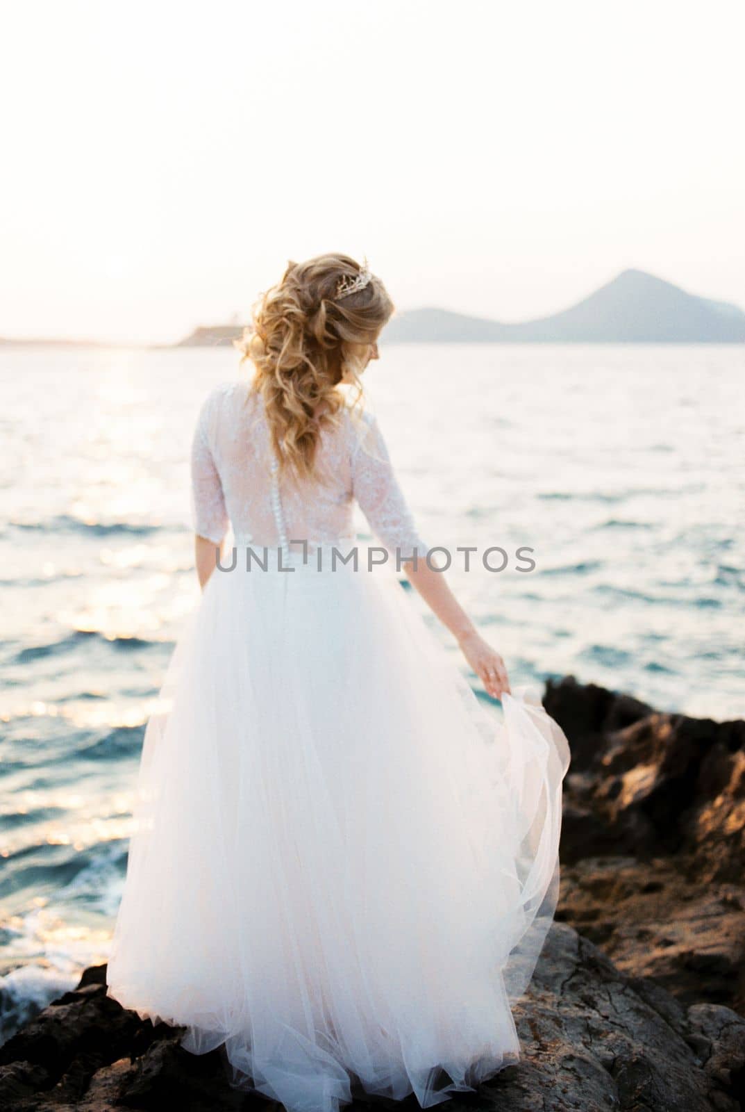 Bride holds the hem of a layered dress on the rocks by the sea. Back view. High quality photo