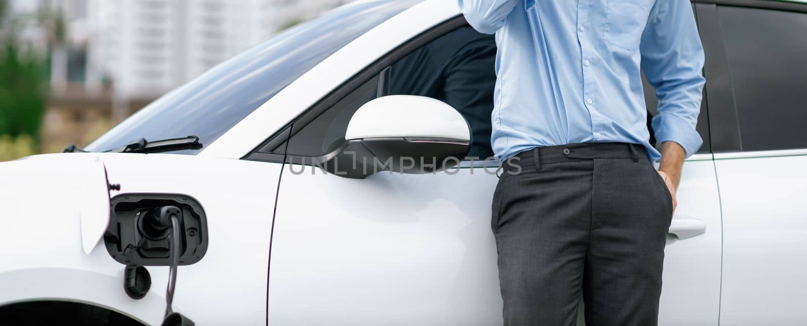 Closeup progressive suit-clad businessman with his electric vehicle recharge his car on public charging station in modern city with power cable plug and renewable energy-powered electric vehicle.