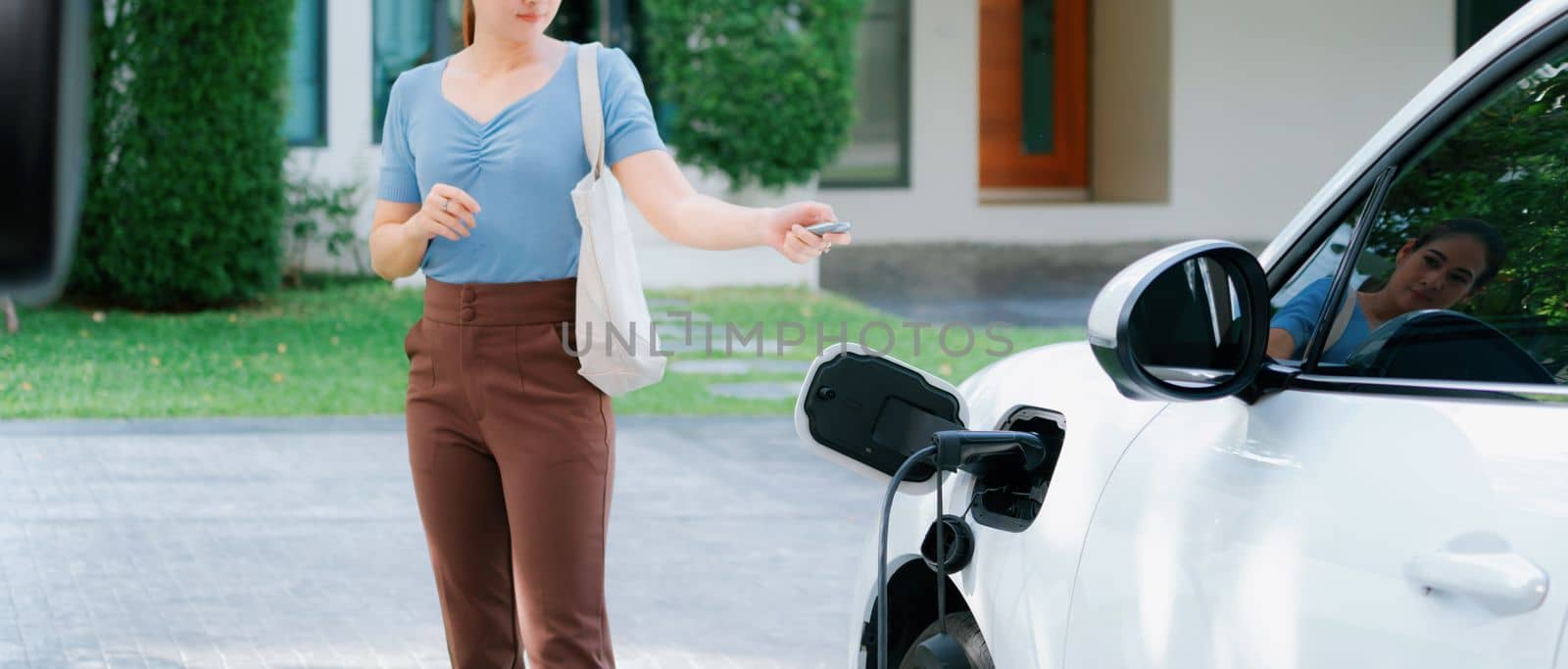 Asian woman lock electric vehicle with remote car key at her garage, EV car recharge battery at home charging station. Alternative clean energy applied in daily life as progressive lifestyle concept.