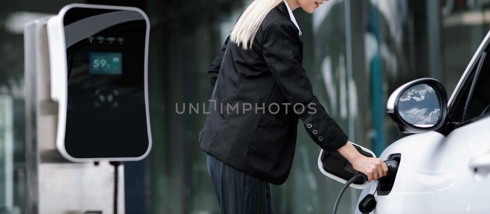 Closeup progressive suit-clad businesswoman with her electric vehicle recharge her car on public charging station in modern city with power cable plug and renewable energy-powered electric vehicle.