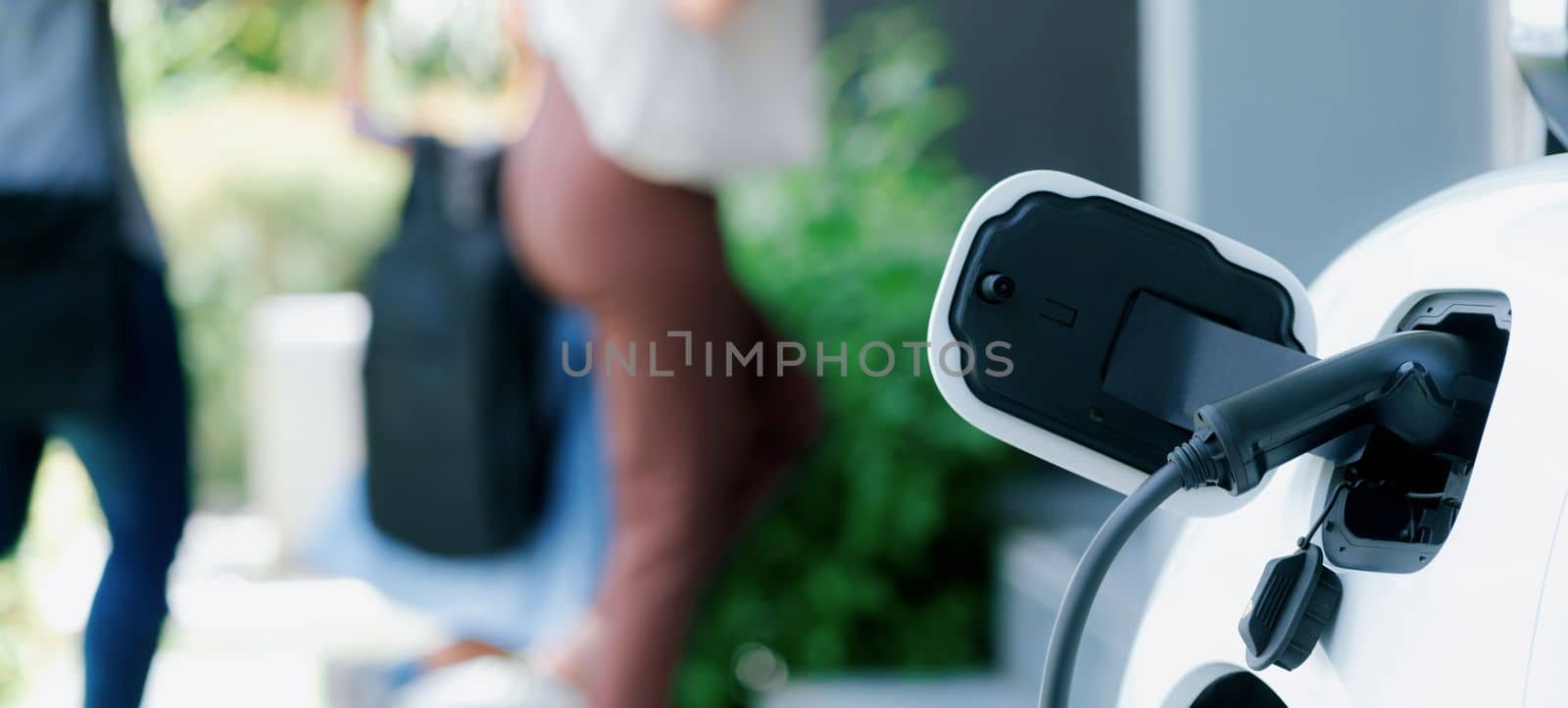 Focus closeup electric vehicle recharging battery from home electric charging station with blurred family in background. Renewable clean energy car for progressive eco awareness lifestyle concept.