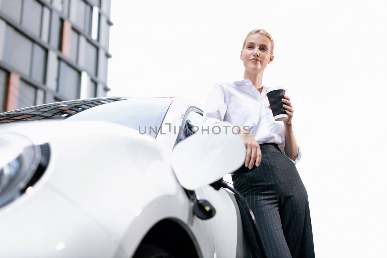 Businesswoman drinking coffee, leaning on electric vehicle recharging at public charging station with residential apartment condos building in background as progressive lifestyle by eco-friendly car.