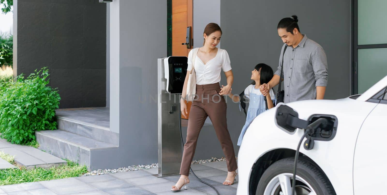Progressive young parents and daughter with electric vehicle and home charging station. Green and clean energy from electric vehicles for healthy environment. Eco power from renewable source at home.