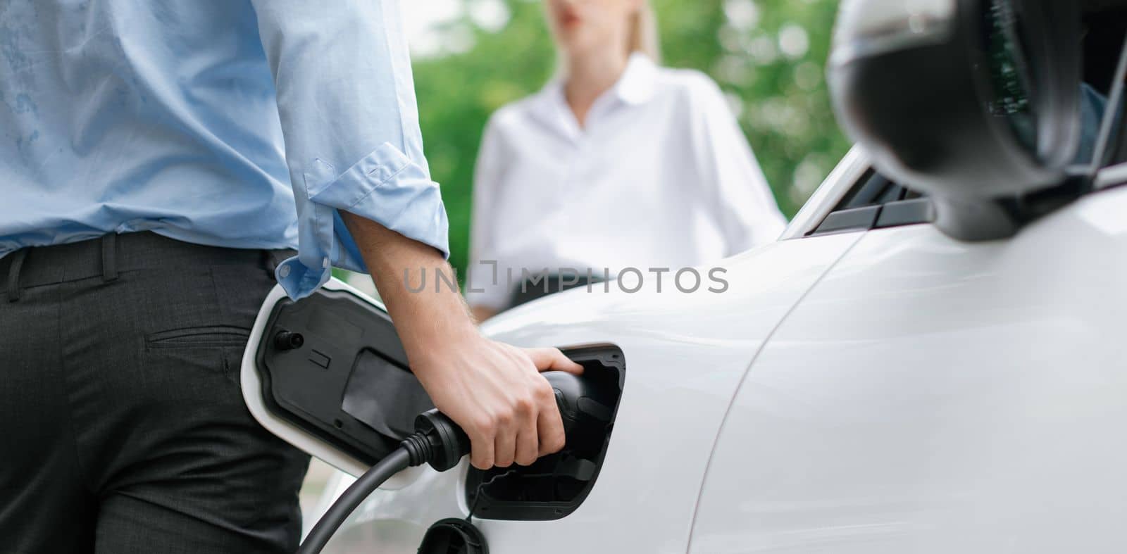 Closeup focus hand insert EV charger to electric vehicle at public charging point in car park with blur business people in backdrop, eco-friendly lifestyle by rechargeable car for progressive concept.