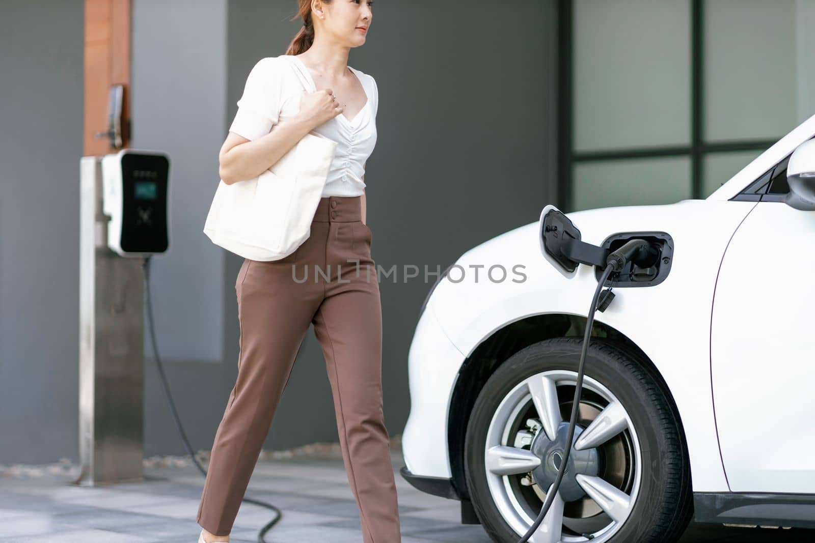 Progressive asian woman and electric car with home charging station. Concept of the use of electric vehicles in a progressive lifestyle contributes to a clean and healthy environment.