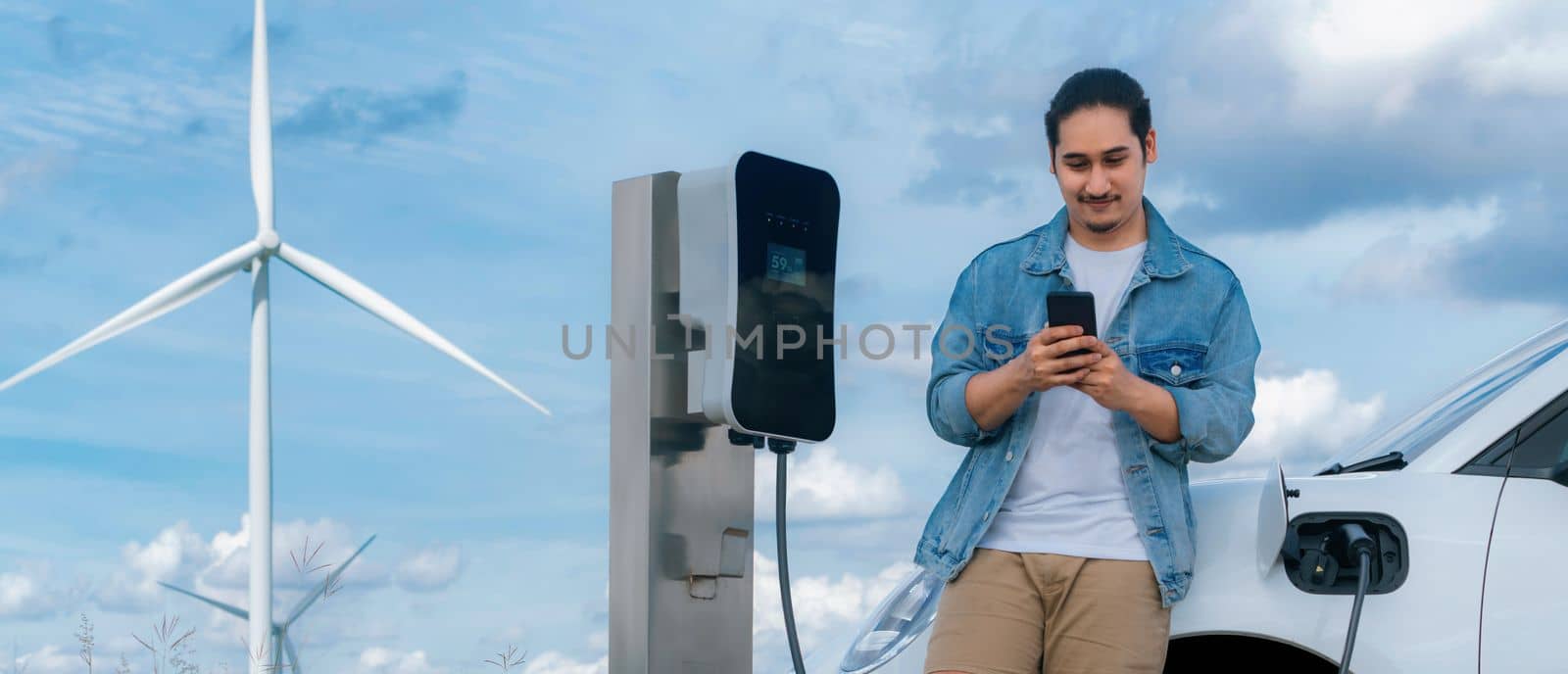 Progressive man with his electric car, EV car recharging energy from charging station on green field with wind turbine as concept of future sustainable energy. Electric vehicle with energy generator.