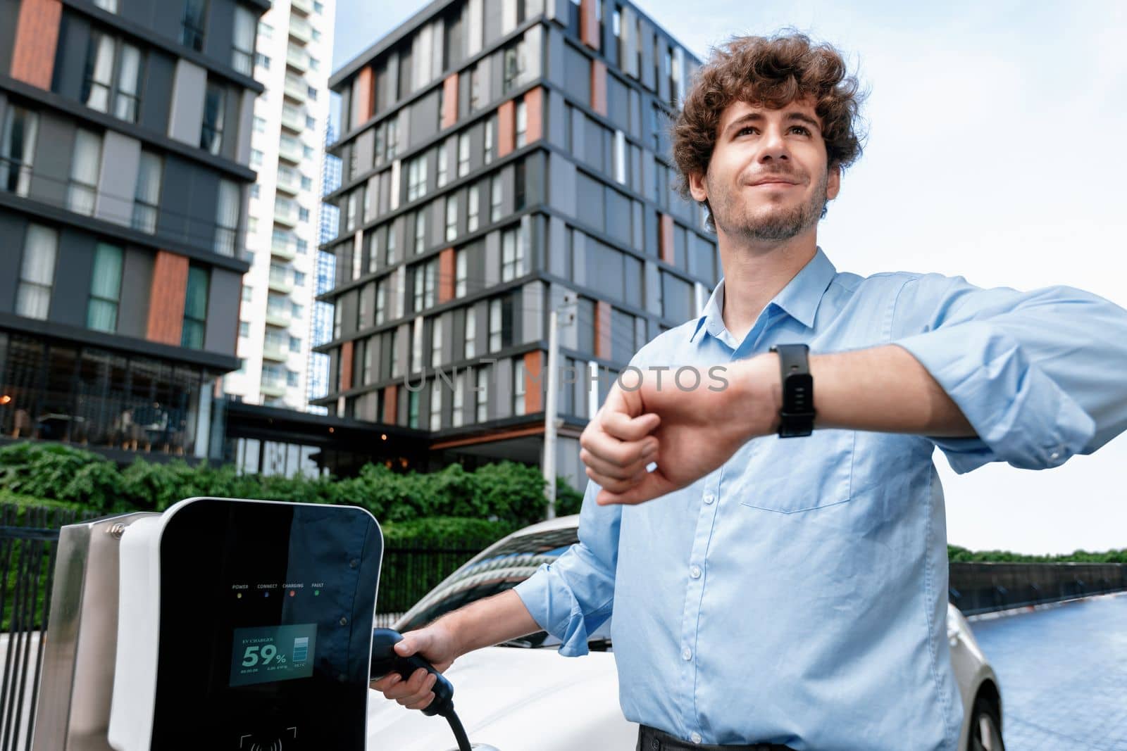 Businessman with smartwatch at modern charging station for electric vehicle with background of residential buildings as concept for progressive lifestyle of using eco-friendly as alternative energy.