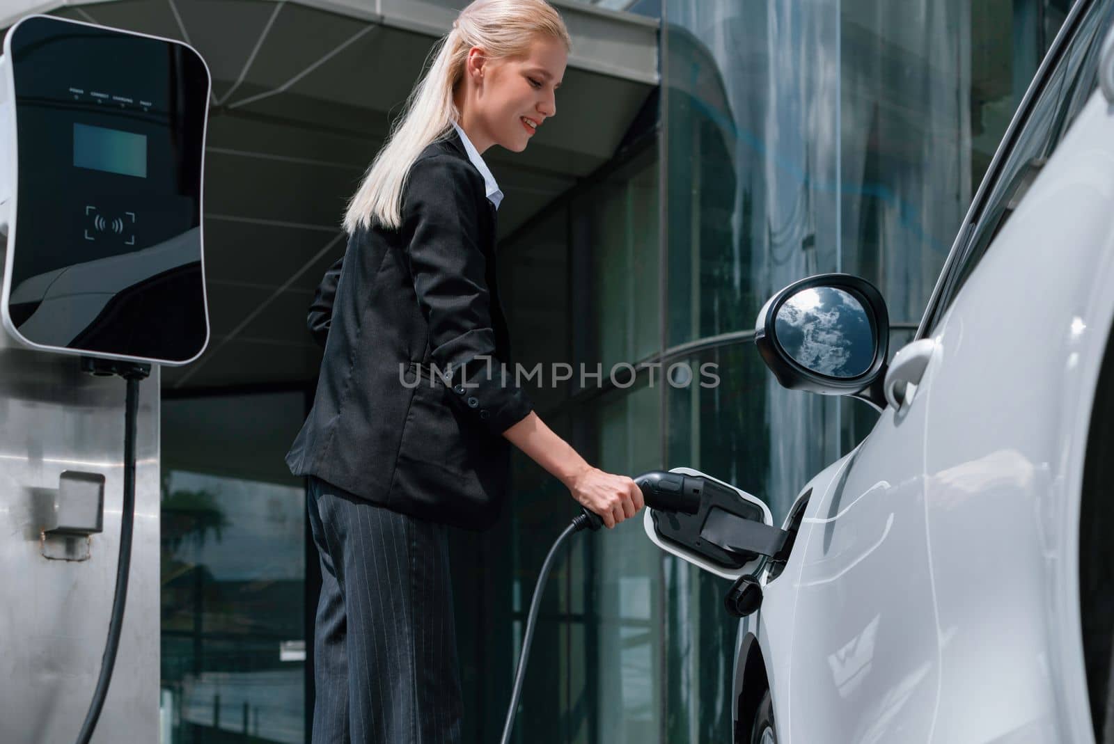 Progressive businesswoman insert charger plug from charging station to her electric vehicle with apartment condo building in background. Eco friendly rechargeable car powered by sustainable energy.
