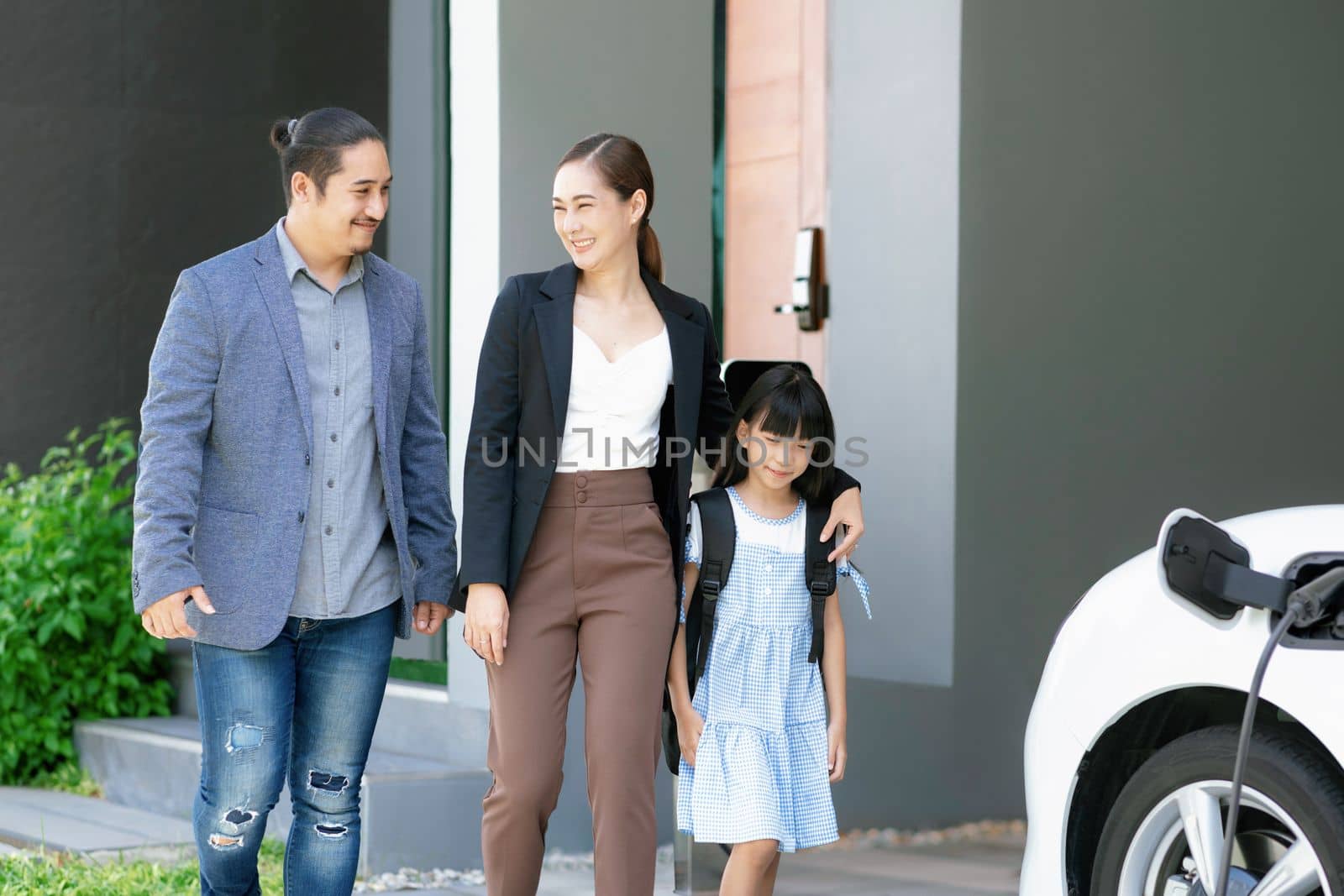 Progressive young parents and daughter with electric vehicle and home charging station. Green and clean energy from electric vehicles for healthy environment. Eco power from renewable source at home.