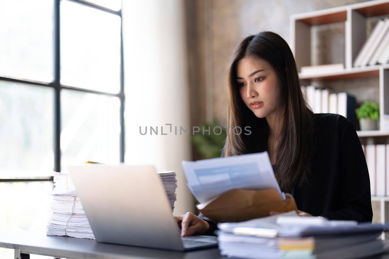 Concentrated young beautiful businesswoman working on laptop and in have documents on hand at bright modern office.
