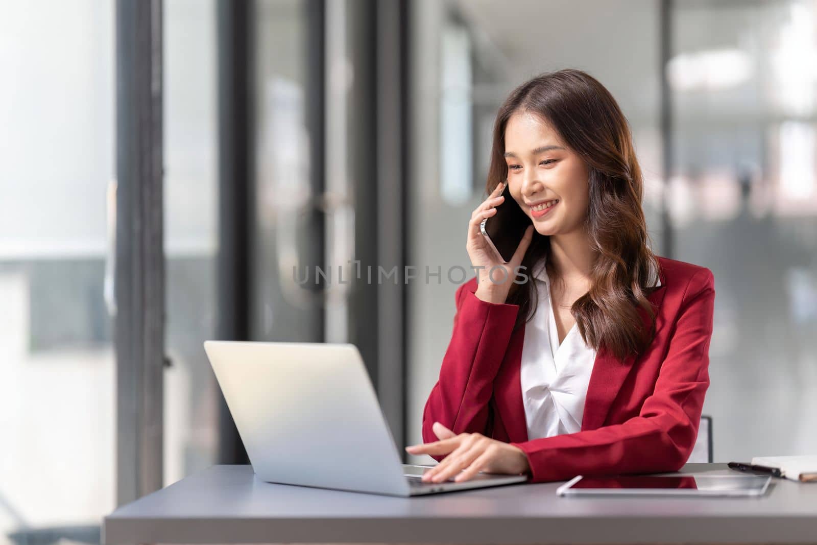 Cheerful young business woman talking on phone working in modern office. Happy positive Asian businesswoman company manager wearing suit making call on cellphone sitting at workplace by nateemee