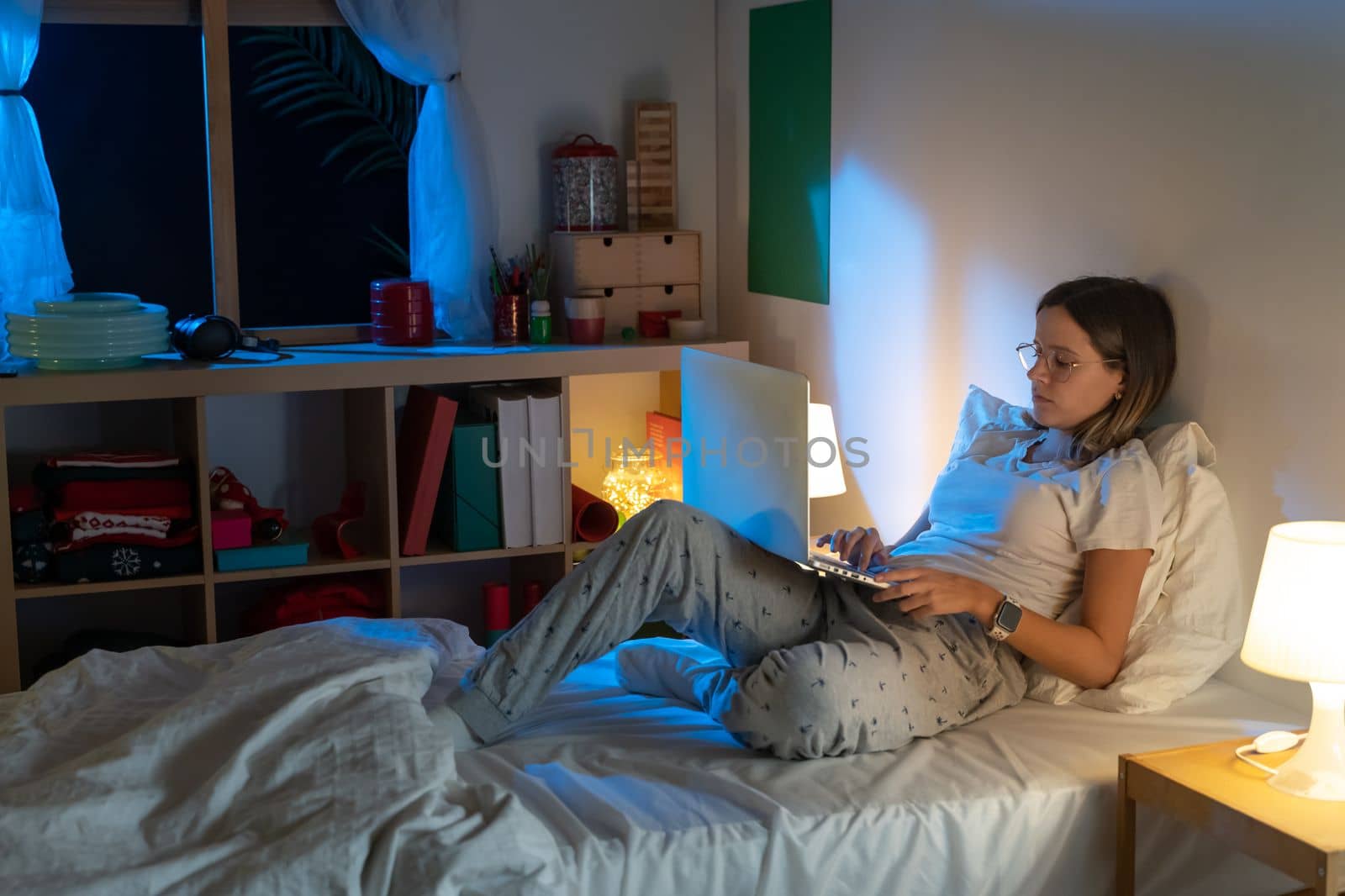 Beautiful young girl sitting on bed at night working with her laptop in the middle of the night. by PaulCarr