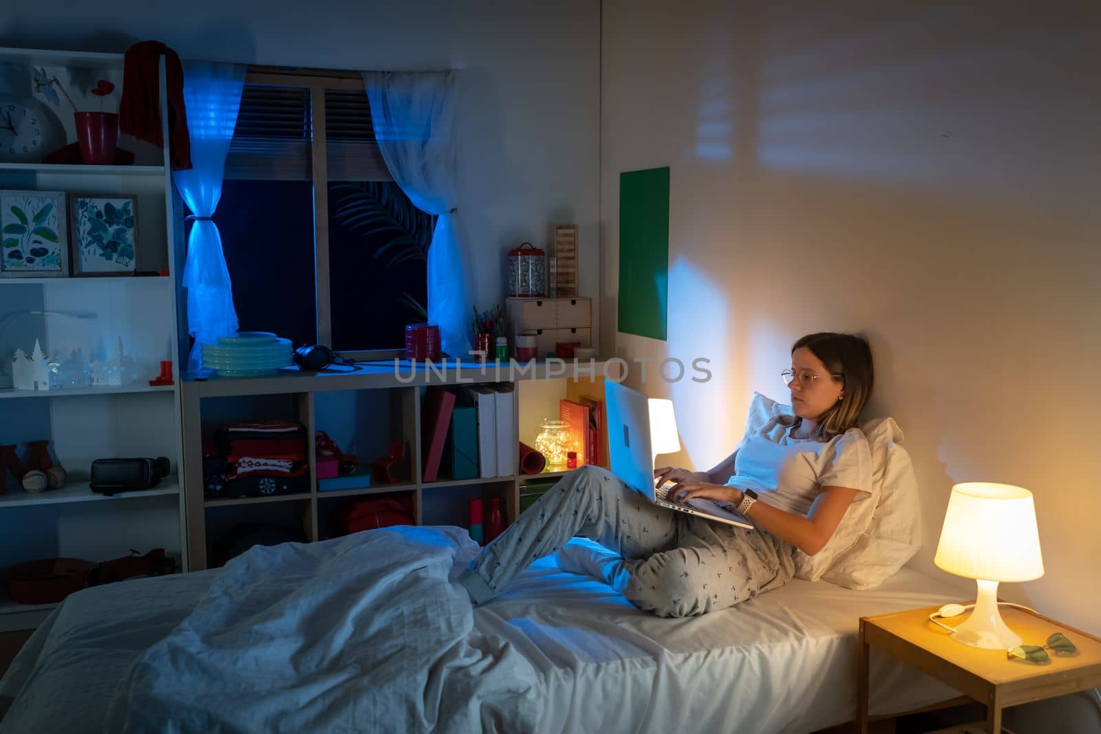 Beautiful young girl sitting on bed at night working with her laptop in the middle of the night. by PaulCarr
