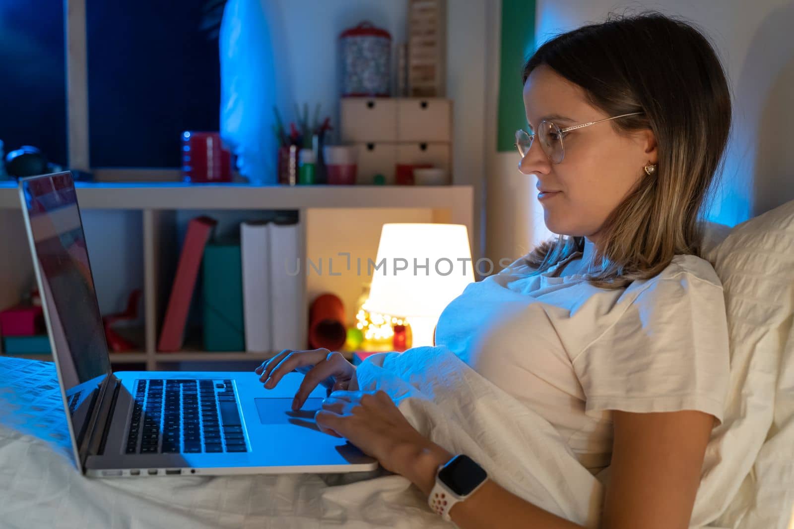 Beautiful young girl sitting on bed at night working with her laptop in the middle of the night. High quality photo