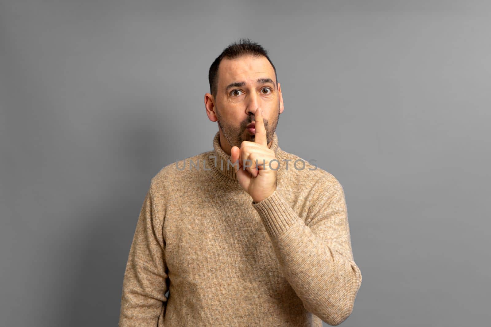 Hispanic handsome man wearing beige turtleneck over isolated gray background asking for silence with finger on lips. Silence and secret concept.