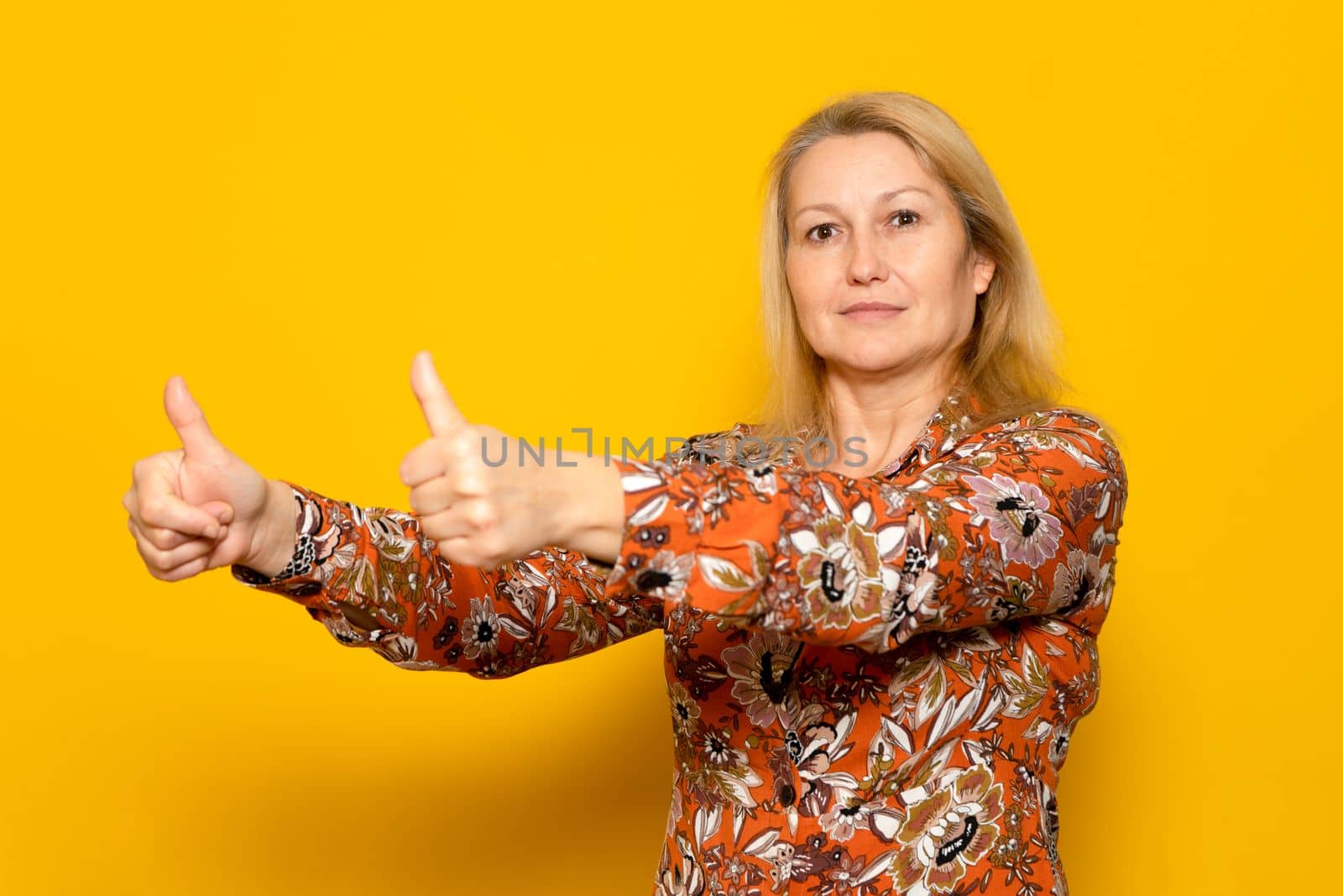 Caucasian woman in her 40s wearing a patterned dress showing thumbs up in agreement, isolated over yellow background