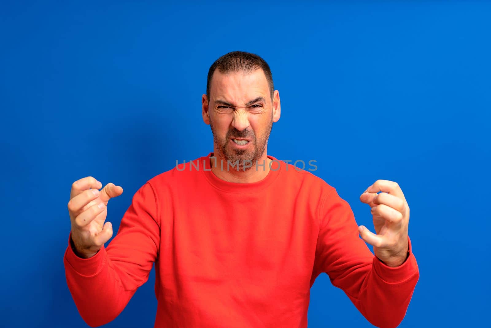 Hispanic man with beard standing over blue isolated background in aggressive and furious attitude with raised hands in the shape of claws