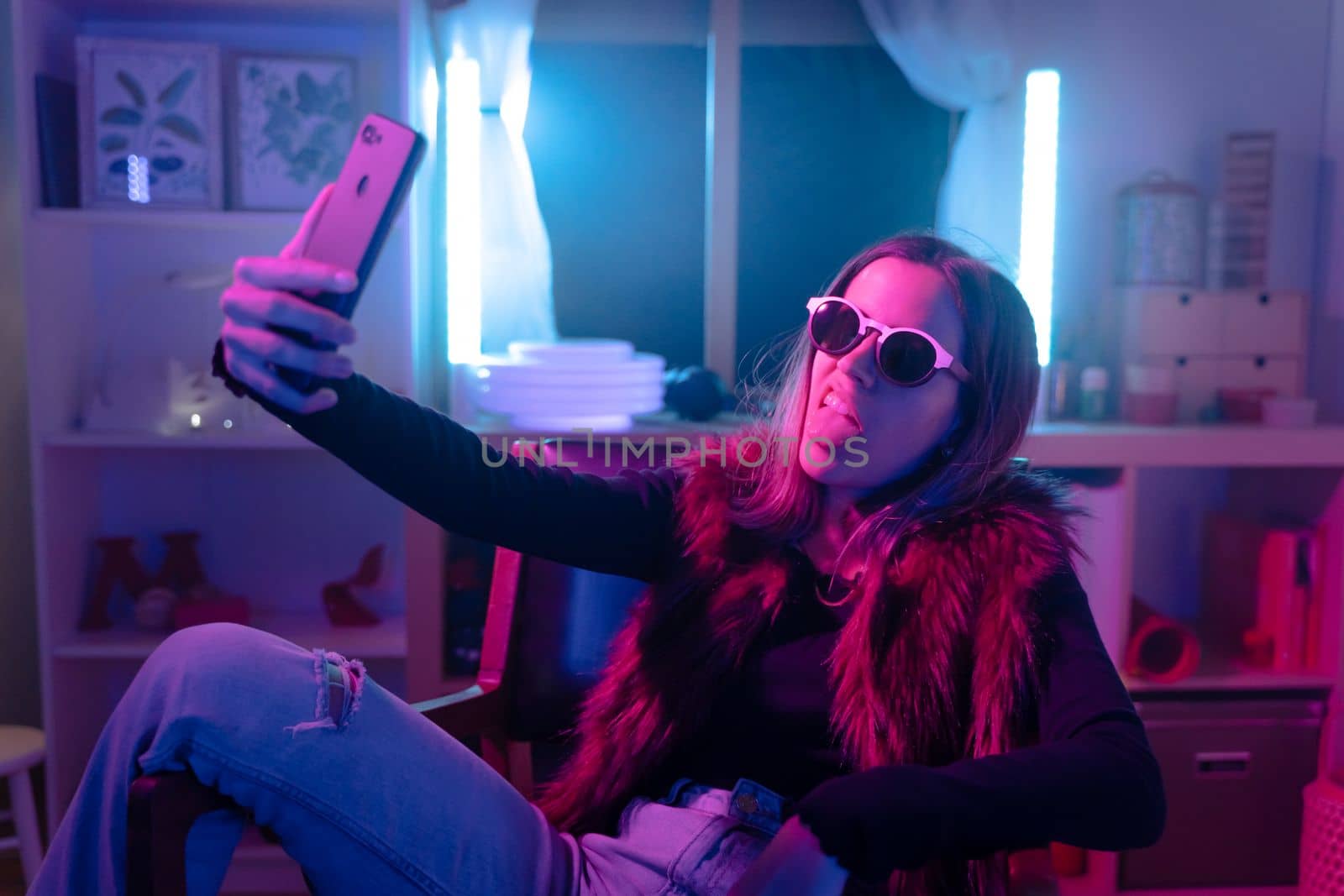 Beautiful young girl taking selfie with her phone on bed with neon colors room. Technology at bed concept. by PaulCarr