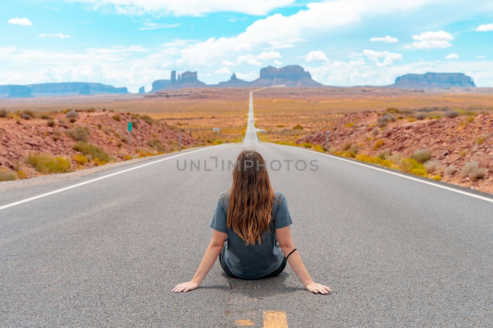 Attractive young woman from back view sitting in the middle of American straight road in the desert. by PaulCarr