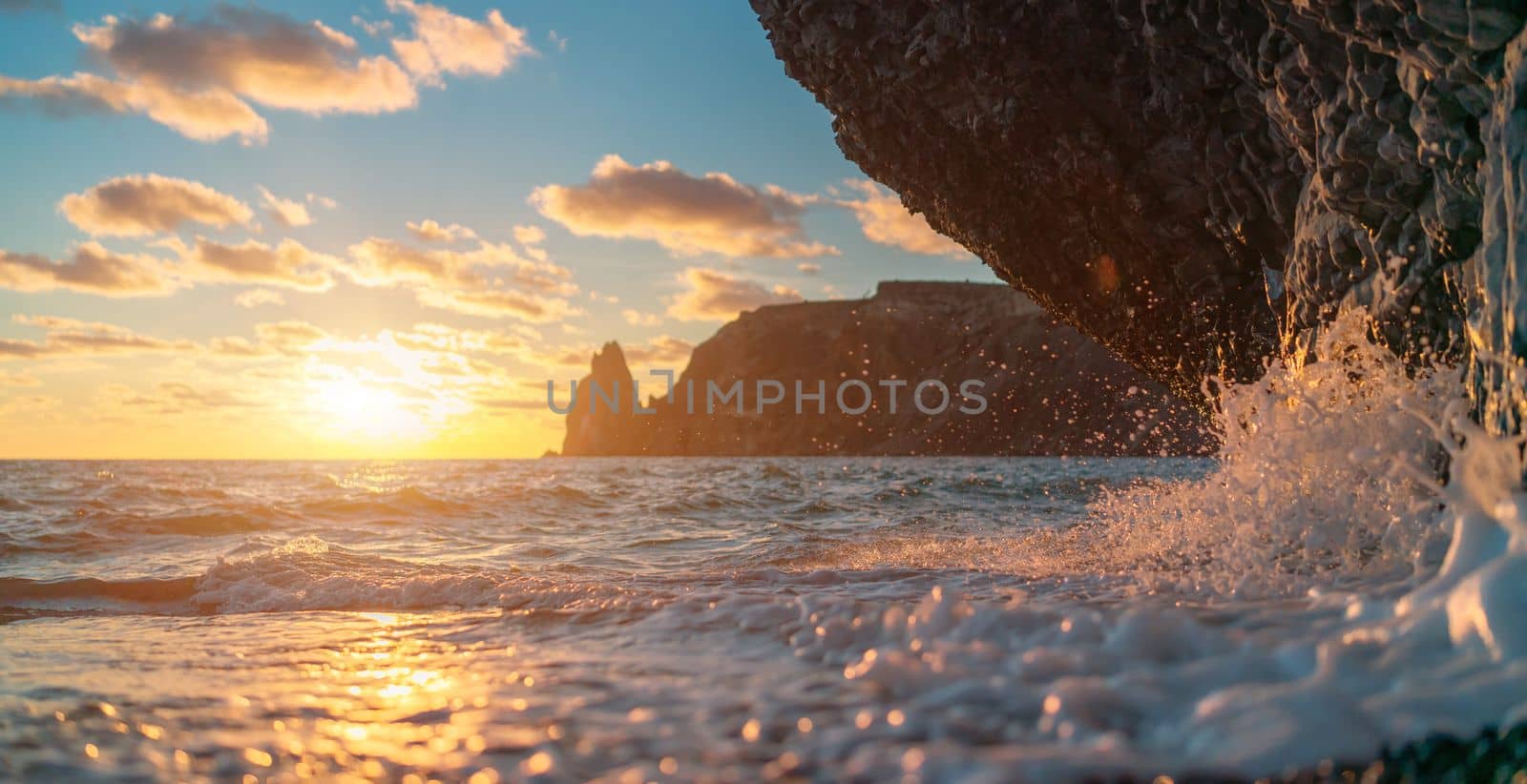 Sea sunset waves with foam. Waves run on pebbles and rock from volcanic basalt. Water breaks into splashes and white foam. Against the background of the cape and the sunset sky