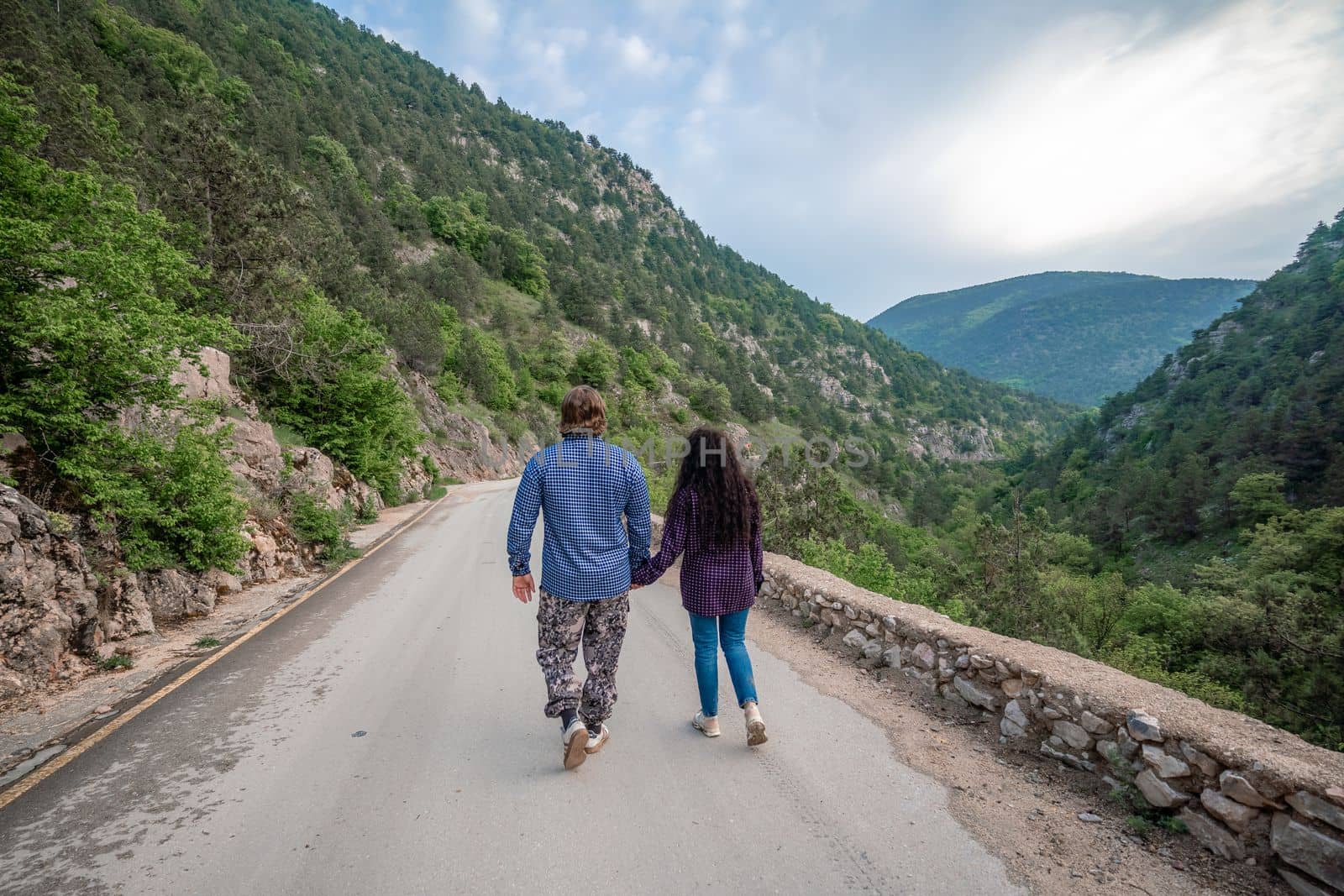A couple in the mountains rejoice in their journey. They hold hands, stand with their backs to the audience, walk along the road and look at the mountains