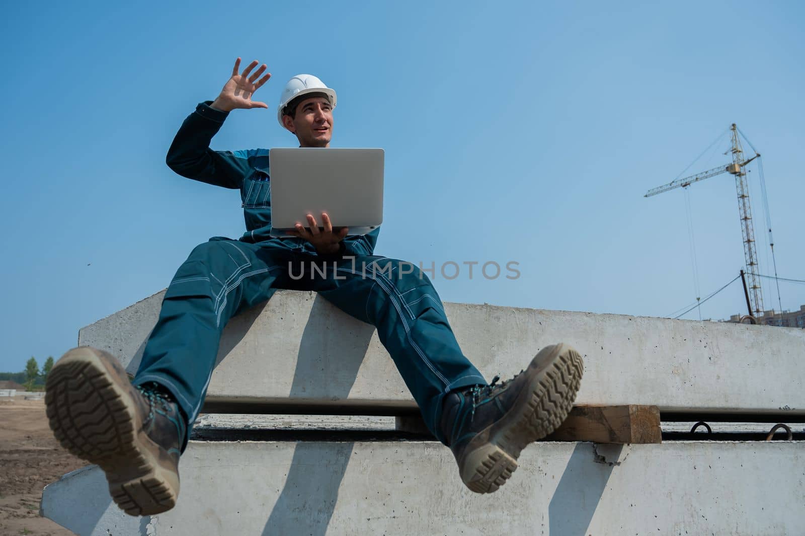 Caucasian male builder in hardhat sits on floor slabs and uses laptop at construction site. by mrwed54