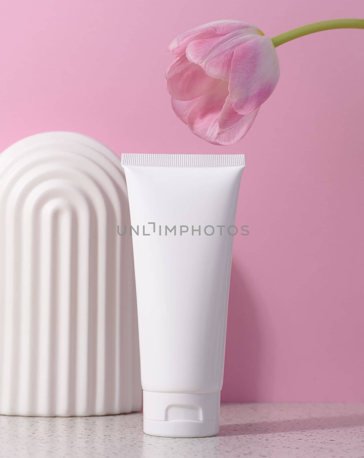 White plastic tube for cosmetic products on a pink background with an arch and a pink tulip, advertising and branding of products