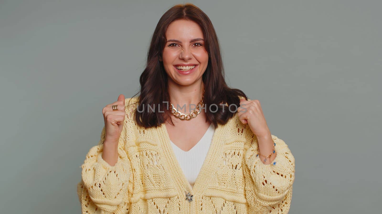 Happy joyful pretty woman in cardigan shouting, raising fists in gesture I did it, celebrating success, winning, birthday, lottery jackpot goal achievemen. Adult girl indoors on gray studio background