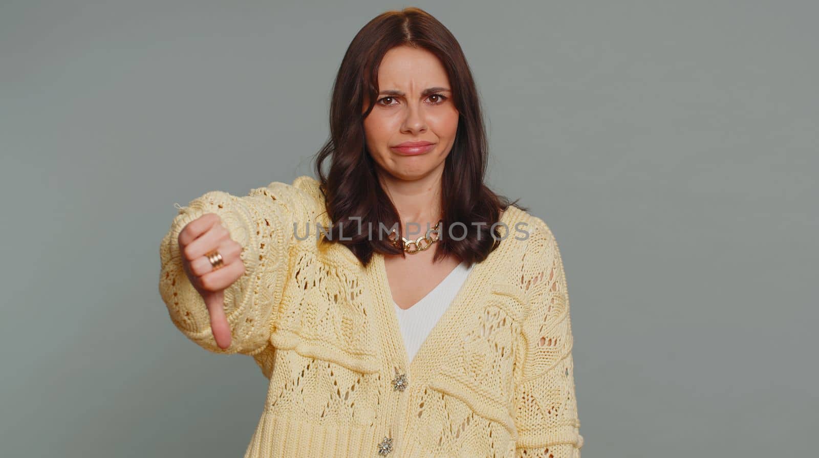 Dislike. Upset unhappy pretty woman in cardigan showing thumbs down sign gesture, expressing discontent, disapproval, dissatisfied, dislike. Young adult girl. Indoor studio shot on gray background