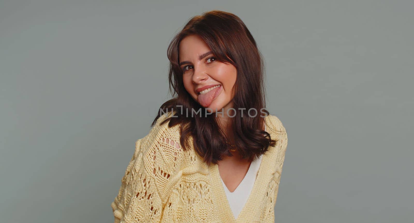 Funny joyful sincere woman 20 years old in cardigan making playful silly facial expressions and grimacing, fooling around showing tongue. Young lovely pretty girl isolated alone on gray background