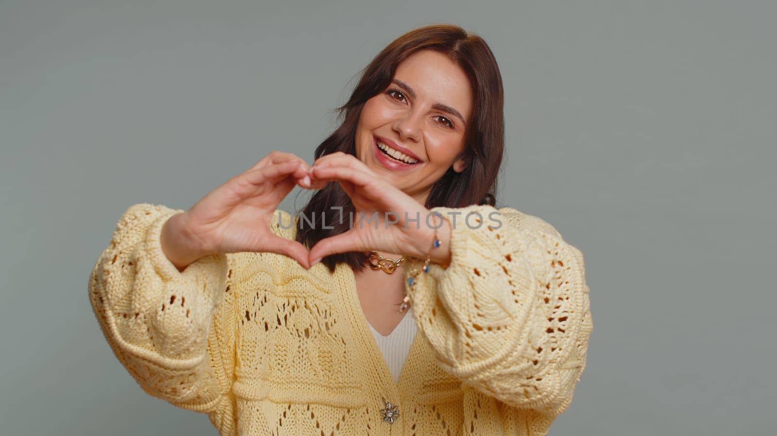 Woman in love. Smiling pretty woman 20s in cardigan use makes heart gesture demonstrates love sign expresses good feelings and sympathy. Young one adult girl isolated alone on gray studio background